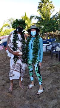 Two individuals stand together wearing traditional Hawaiian outfits and leis amidst a beach setting.