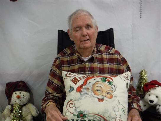 Elderly man holds a Christmas pillow while sitting near decorative winter items in a cozy setting.
