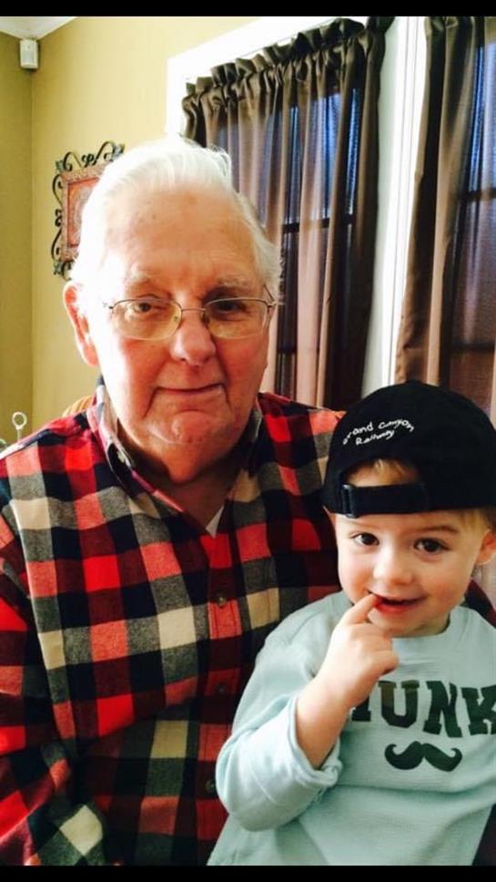 An elderly man wearing glasses and a plaid shirt holds a young boy in a cap, both smiling.