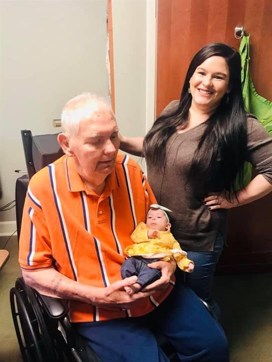 Elderly man sitting in a wheelchair holds a doll while a young woman stands proudly beside him.