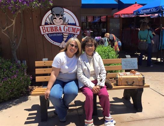 Two friends are sitting on a bench outside a restaurant on a sunny day, smiling happily.
