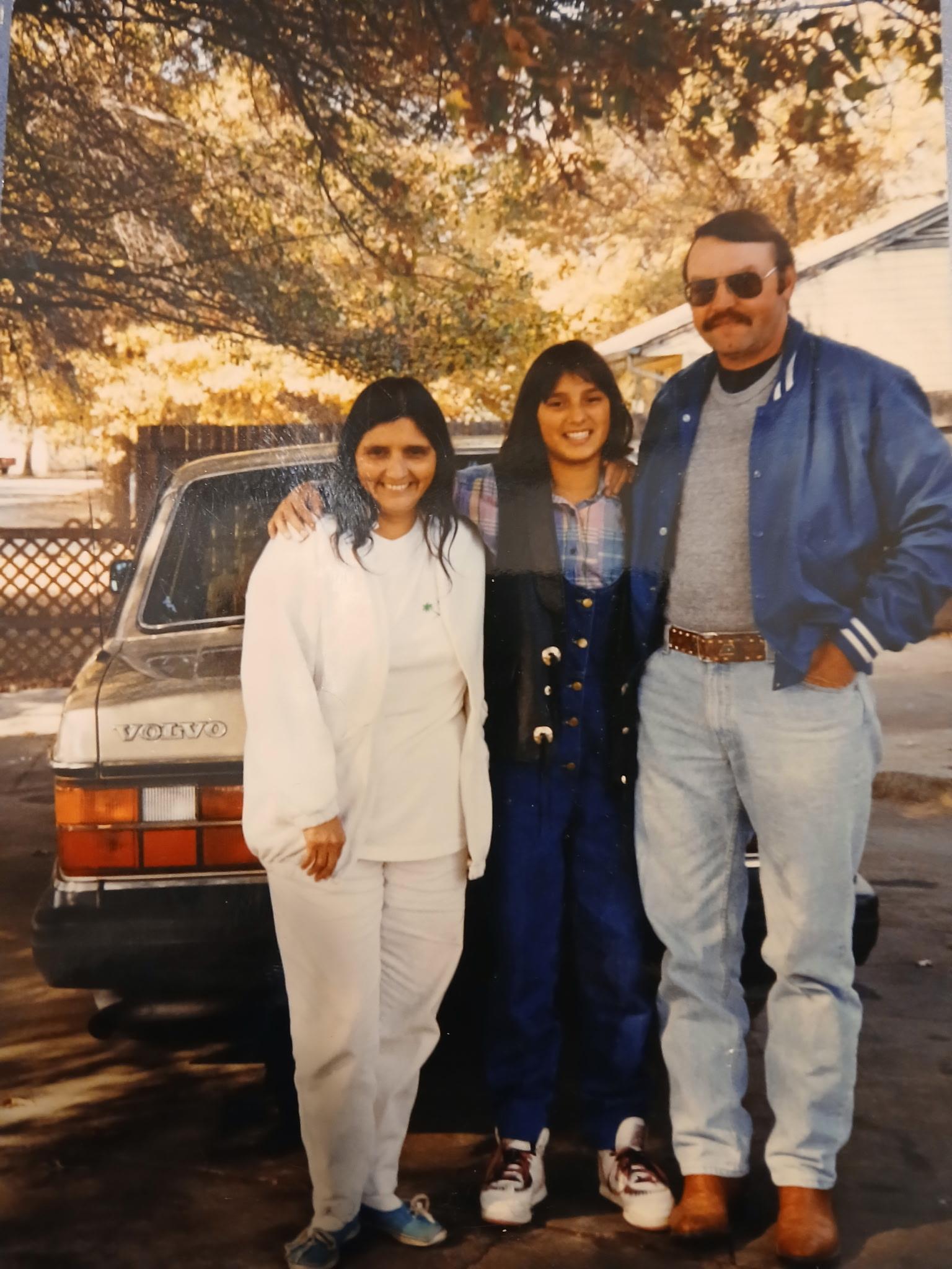 Friends smile in autumn outfits, sharing a cheerful moment by a classic car outdoors.