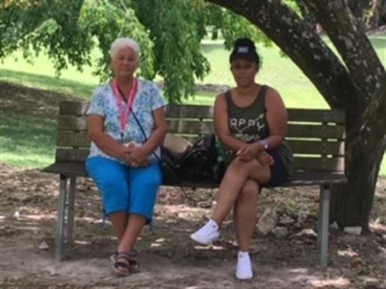 Two women sit comfortably on a park bench, enjoying the shade of a large tree on a sunny day.
