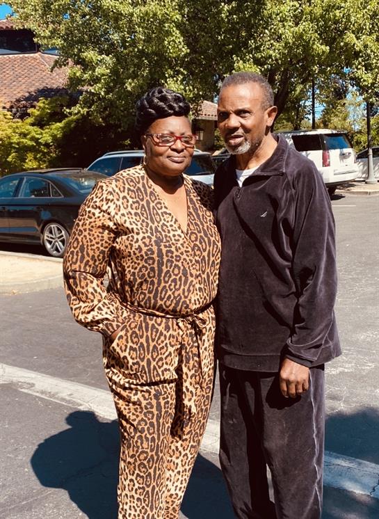 A man and woman stand closely together on a warm afternoon, showcasing their stylish outfits.