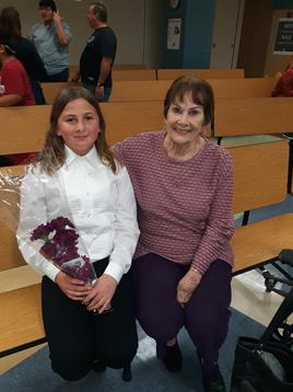 A young girl sits next to her grandmother in an auditorium, holding a bouquet.
