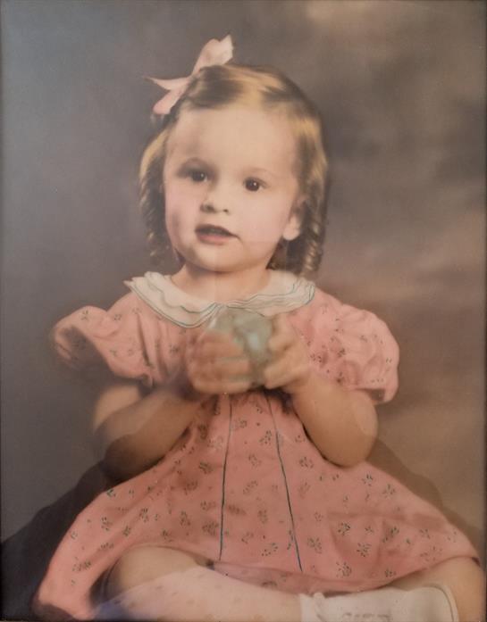 A young girl with blond hair in a pink dress holds a small object, looking thoughtfully.