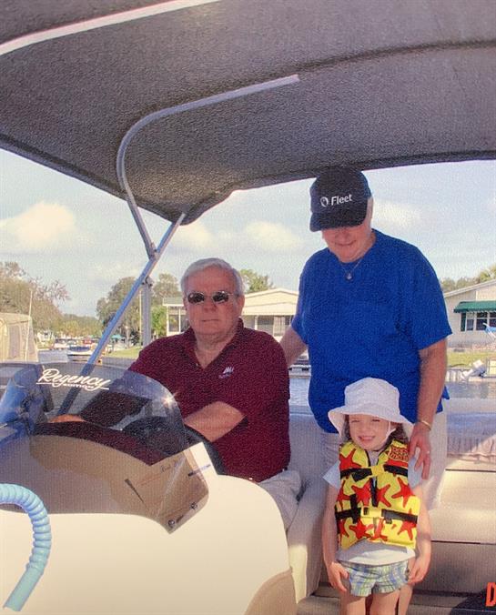 A family is smiling together on a boat, with a child in a colorful shirt, enjoying the warm weather.