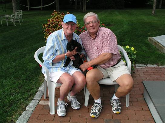 Married couple sits on garden chairs holding a small black cat, smiling on a sunny day.