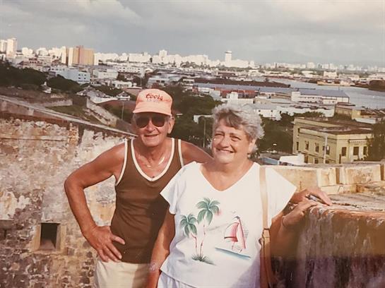 Older couple stands on a historic wall, smiling with a beautiful view of San Juan's cityscape.