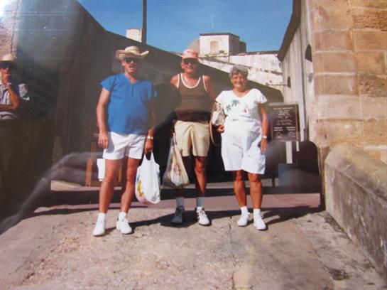Three people stand together in a sunlit street, dressed for summer and smiling cheerfully.