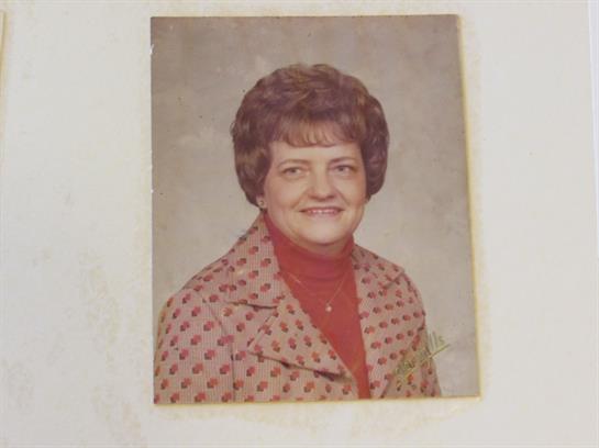 A smiling elderly woman dressed in a patterned coat stands against a neutral backdrop.