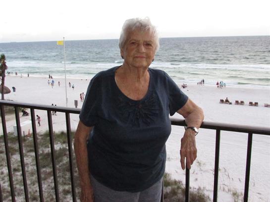 Senior woman stands on a balcony overlooking a serene beach with gentle waves and visitors.