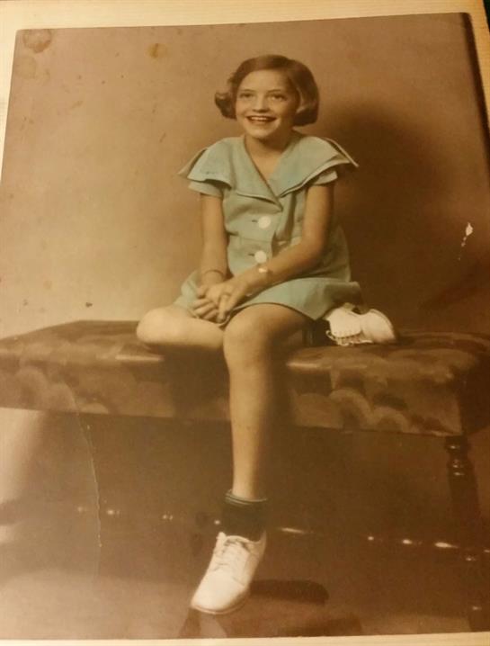 A young girl in a light blue vintage outfit smiles happily on a wooden bench.
