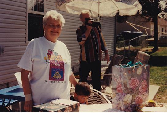 A grandmother stands beside a table at a backyard gathering while a child plays below her.