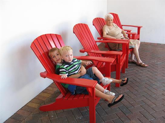Two children sit together in a bright red chair while an older woman observes nearby.