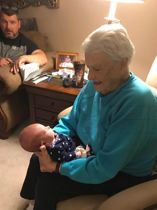 A happy elderly woman holds a baby in a cozy living room with family nearby.