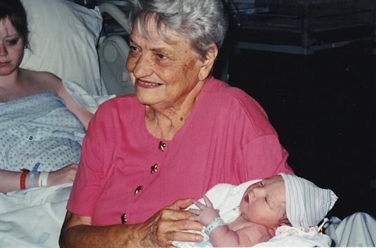 An elderly woman smiles warmly while holding her newborn grandchild in a comfortable setting.
