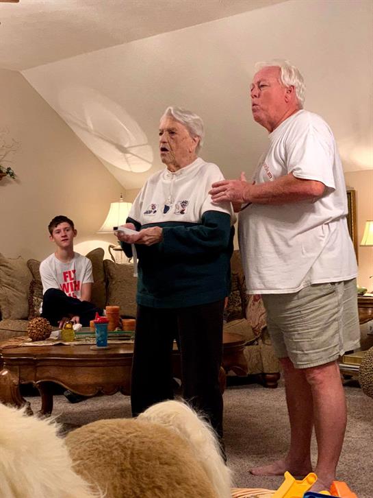 An elderly couple joyfully sings as a younger family member watches in a cozy living room.