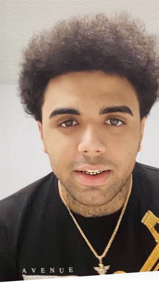 A young man with curly hair smiles confidently while wearing casual attire and jewelry.