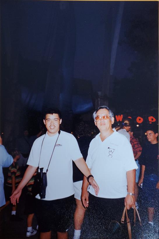 Two men smile and pose together amidst a vibrant outdoor event with festive lights.