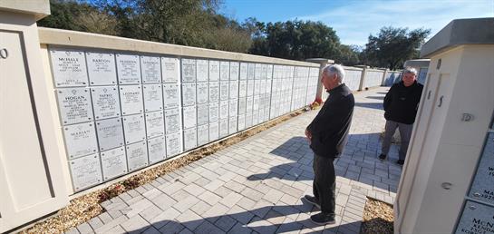 A man stands thoughtfully in front of a memorial wall, reflecting on the names displayed.