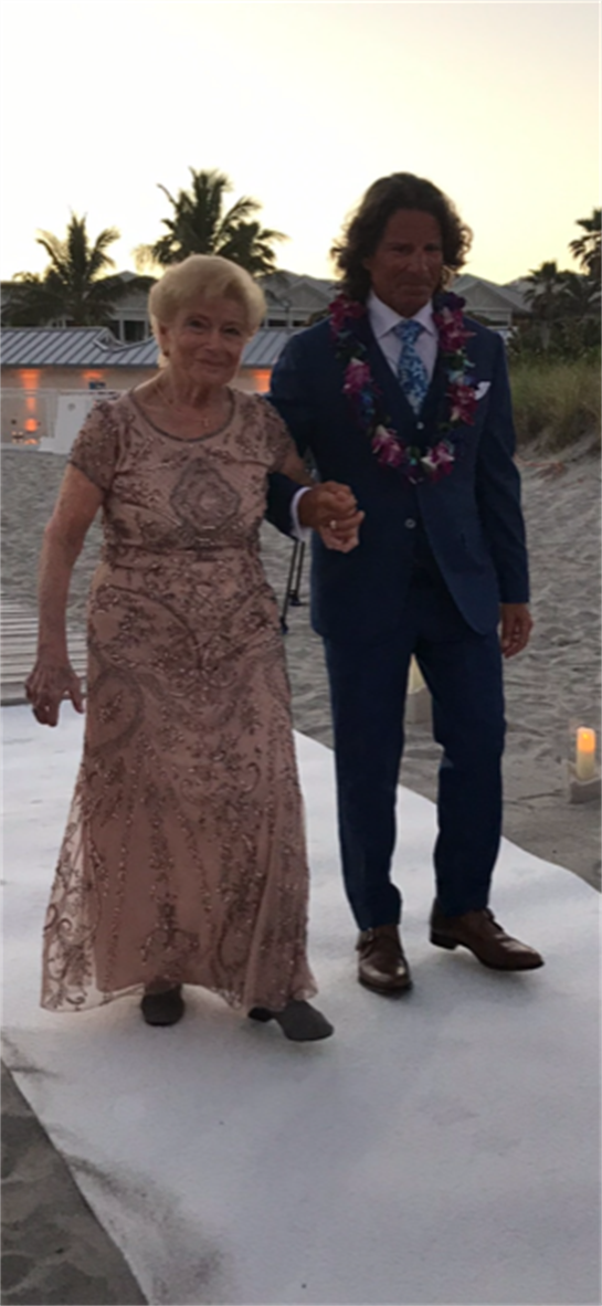A man in formal attire accompanies an elderly woman in a beautiful dress along a beach pathway.