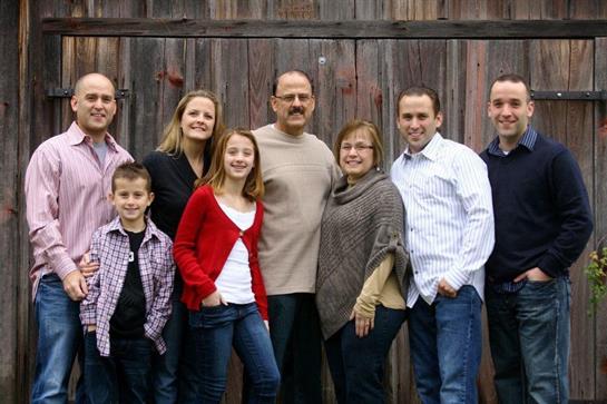 A happy family poses outside in casual clothes, smiling by a wooden backdrop.