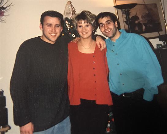 Three friends smile while standing beside a decorated Christmas tree in a cozy indoor setting.