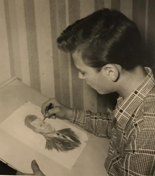 A boy concentrates on creating a detailed portrait at a desk in a warmly lit room.