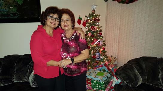 Two women embrace joyfully next to a Christmas tree adorned with colorful decorations and gifts.
