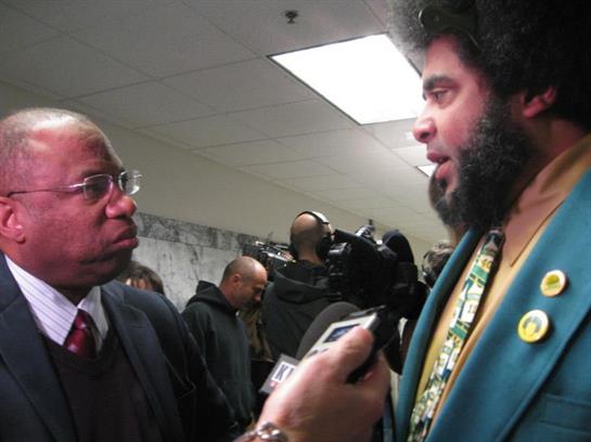 Two men engage in a serious conversation while others gather in the background.