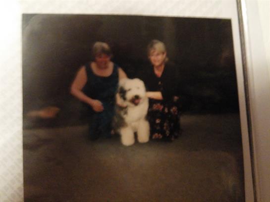 Two women smile as they pose with a fluffy white dog in a warm indoor setting at night.