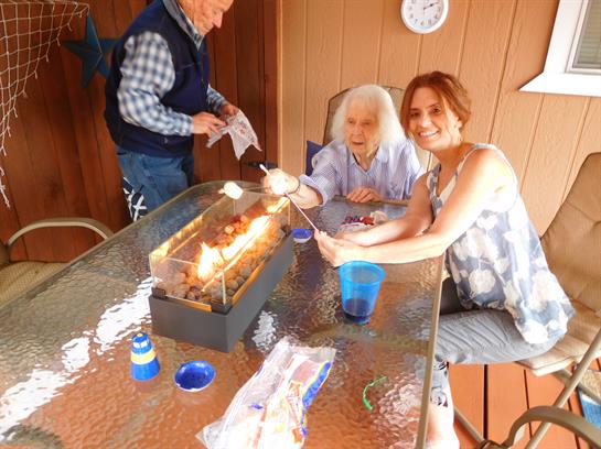 Two women roast marshmallows while an older man oversees the fire in a cozy backyard.