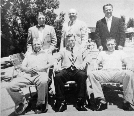 Six men pose together, blending formal and casual wear in the sunlight.