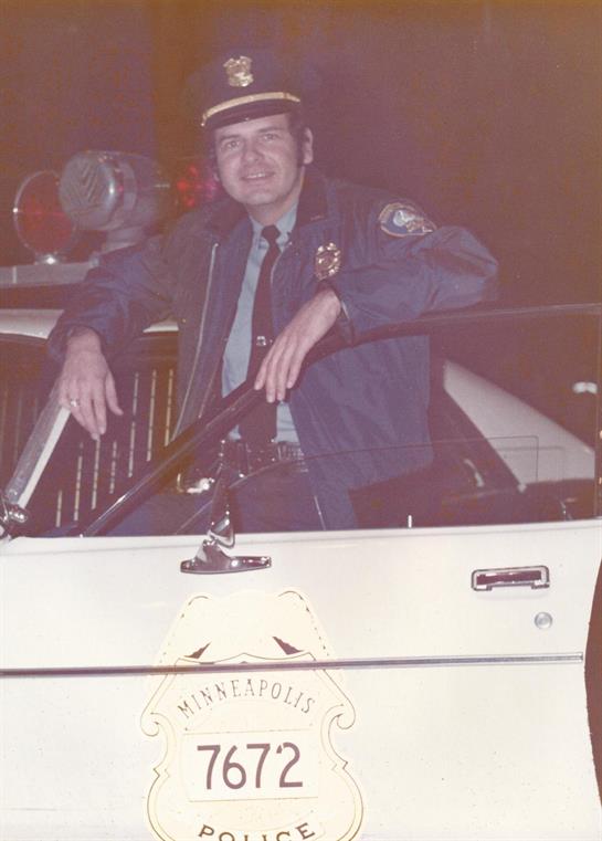 A police officer is casually leaning against a patrol car at night, illuminated by streetlights.