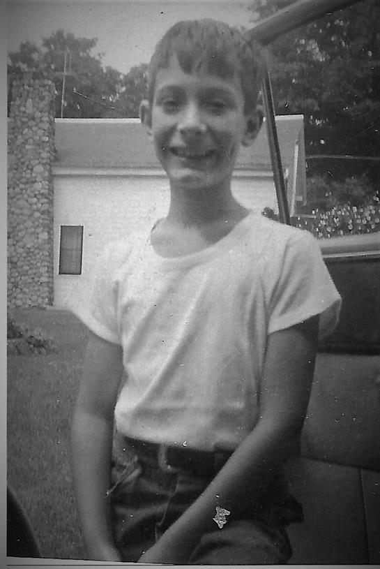 A cheerful young boy with a friendly smile stands beside a car in a sunny neighborhood.