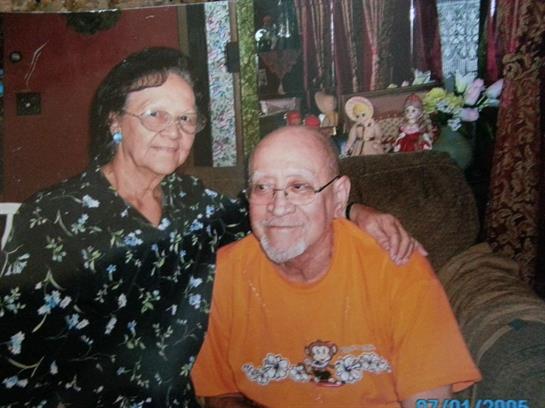 Elderly couple smiles while seated in a comfortable living room filled with personal decor.