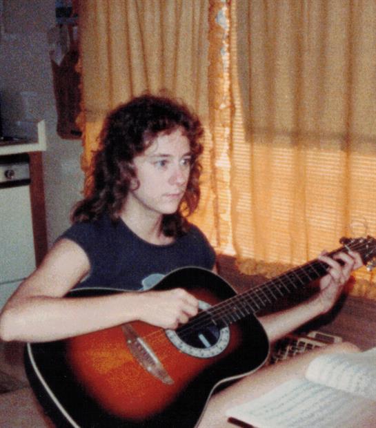 A girl strums an acoustic guitar while sitting in a sunlit room with yellow curtains.