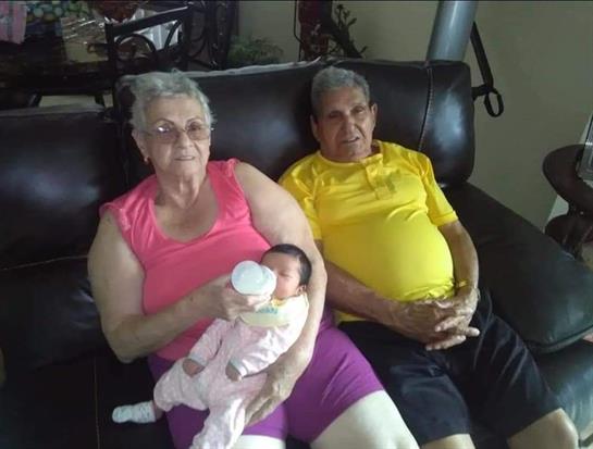 An elderly couple relaxes on a couch, with the grandmother holding a baby. Warm familial moment.