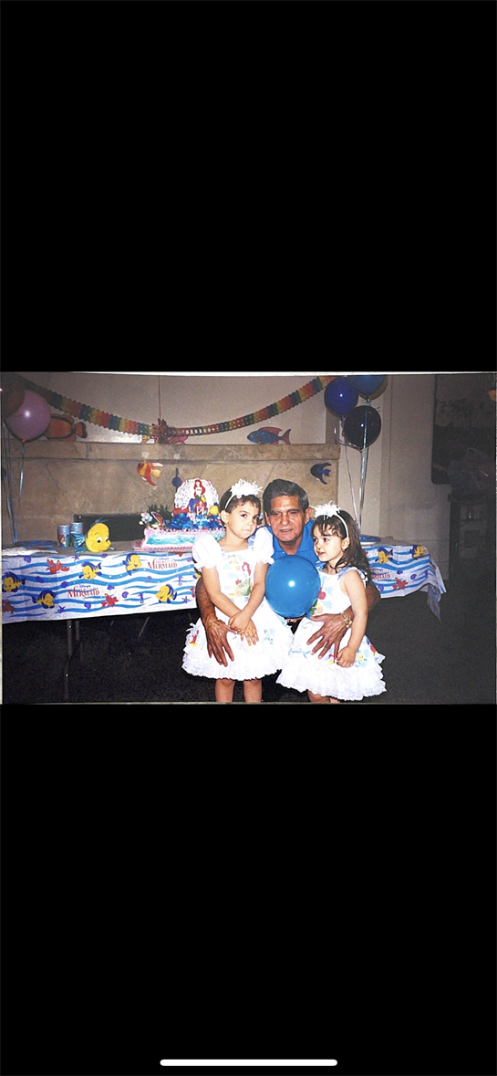 Two girls in white dresses hold balloons while posing with a man at a festive birthday gathering.