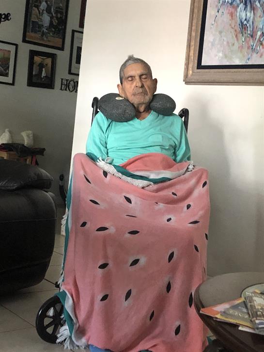 An elderly man sits comfortably in a chair, wrapped in a blanket, with a neck pillow for support.