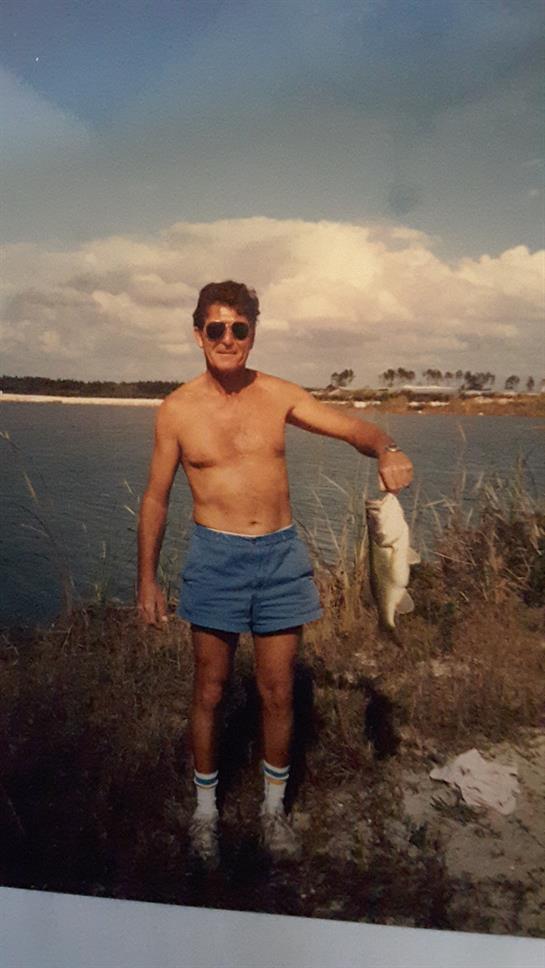 A man in swim trunks and sunglasses holds a big fish by the water under a clear sky.