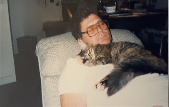 A relaxed man sits on a couch with a sleeping cat on his chest, enjoying a peaceful afternoon.
