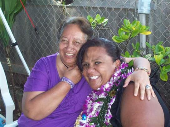 Two women joyfully embrace while wearing flower leis amidst greenery, enjoying a sunny outdoor day.