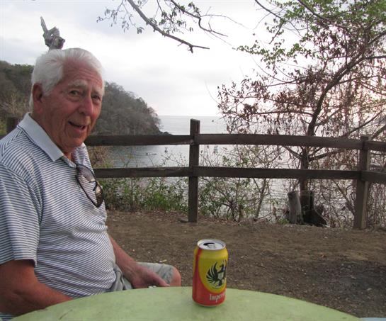 An elderly man sits at a green table, sipping a drink while enjoying the peaceful lakeside view.