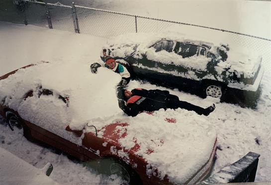 Two people play in the snow on top of cars buried under a thick layer of snow in winter.