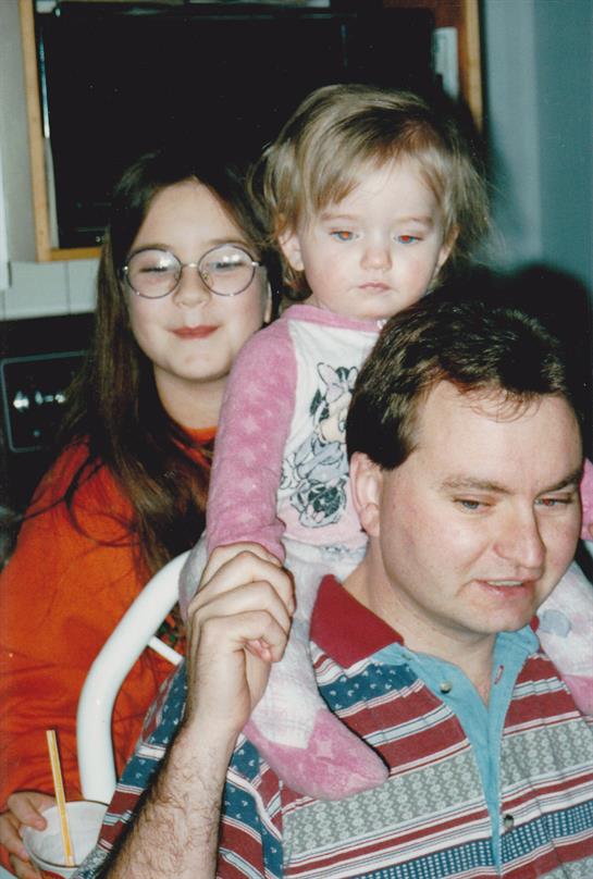 A father holds his daughter on his shoulders while a woman smiles behind them in a cozy home.