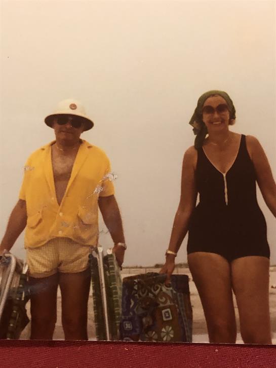 A couple walks hand in hand along the beach with bags, enjoying a sunny summer day.