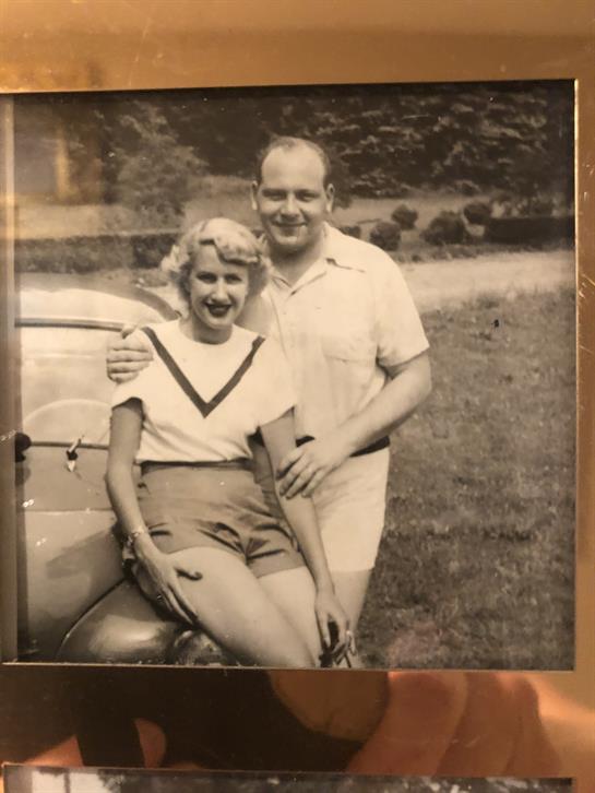 Young couple poses happily together near a vintage car in a sunny outdoor location.
