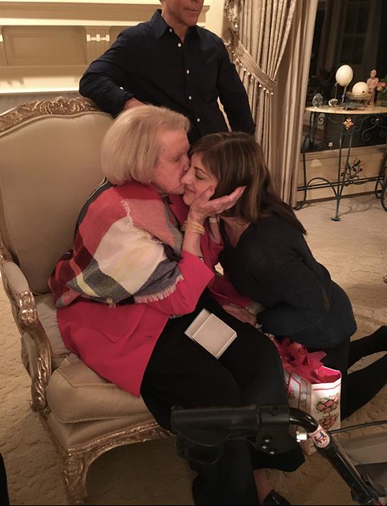 Two women share a heartfelt kiss while seated in an elegant living room during a family gathering.